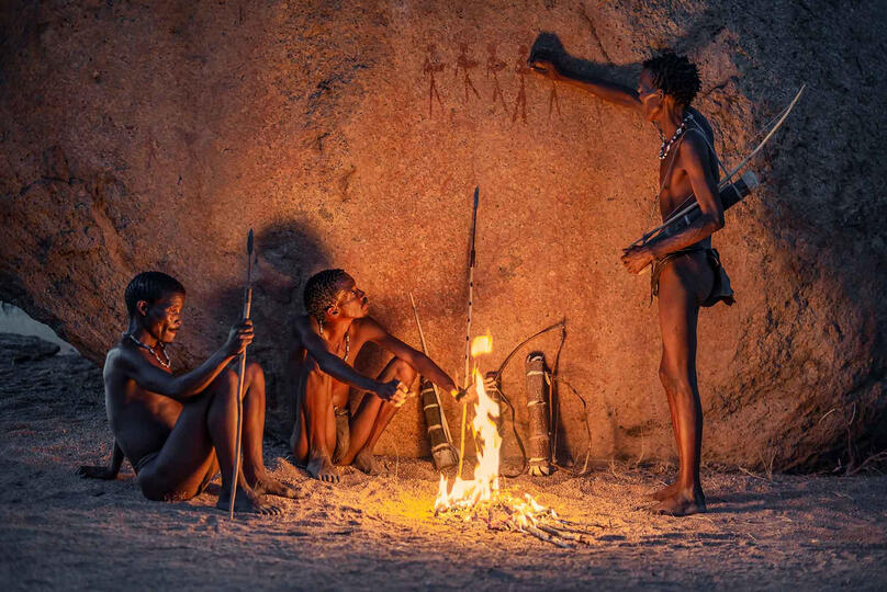 The Fire of the Ancestors Group of San tribesmen resting under an original ancient rock painting left thousands of years ago by their ancestors Buy Print